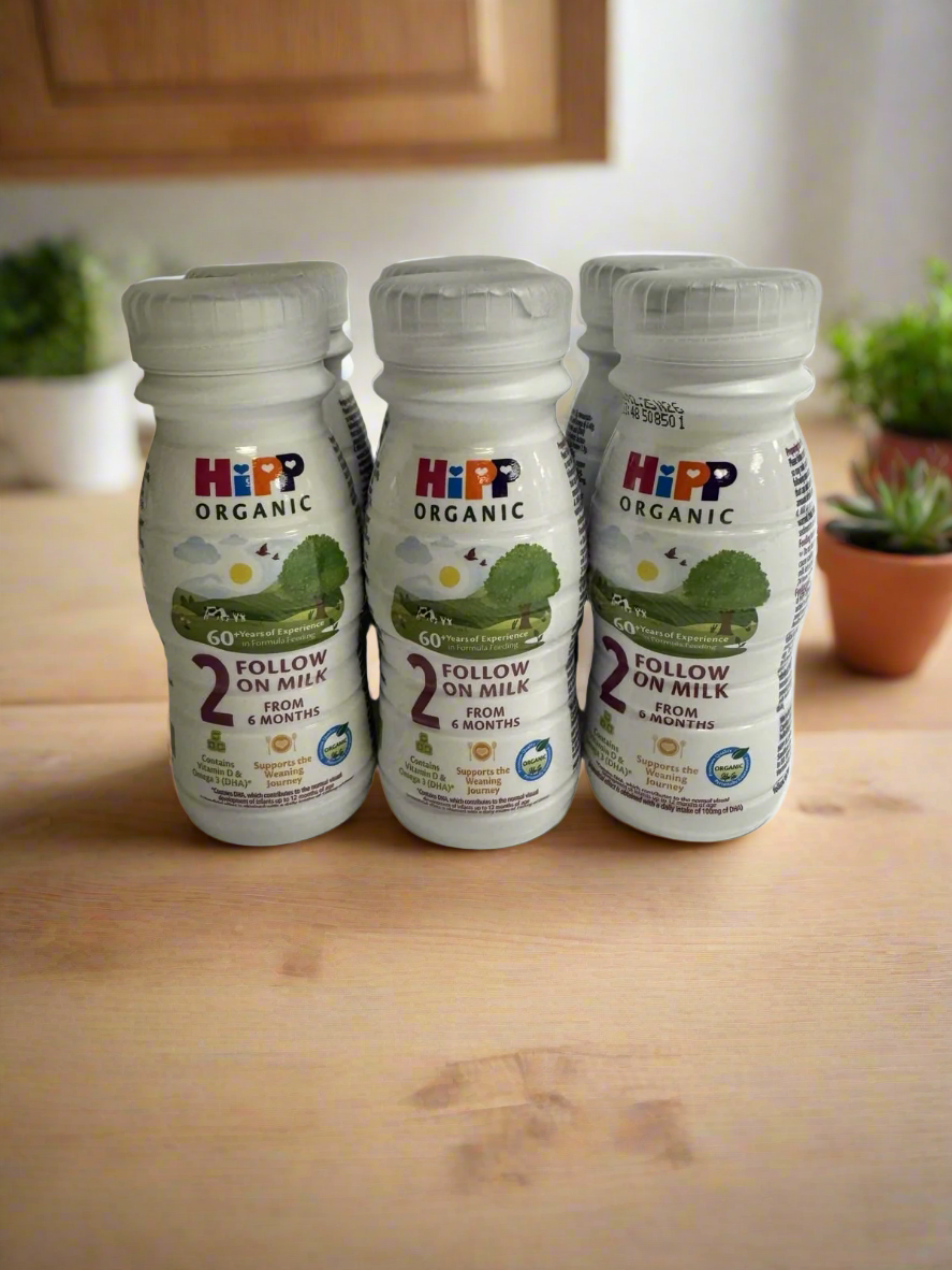 Six HiPP Organic Follow On Milk bottles on a wooden surface with a kitchen background.