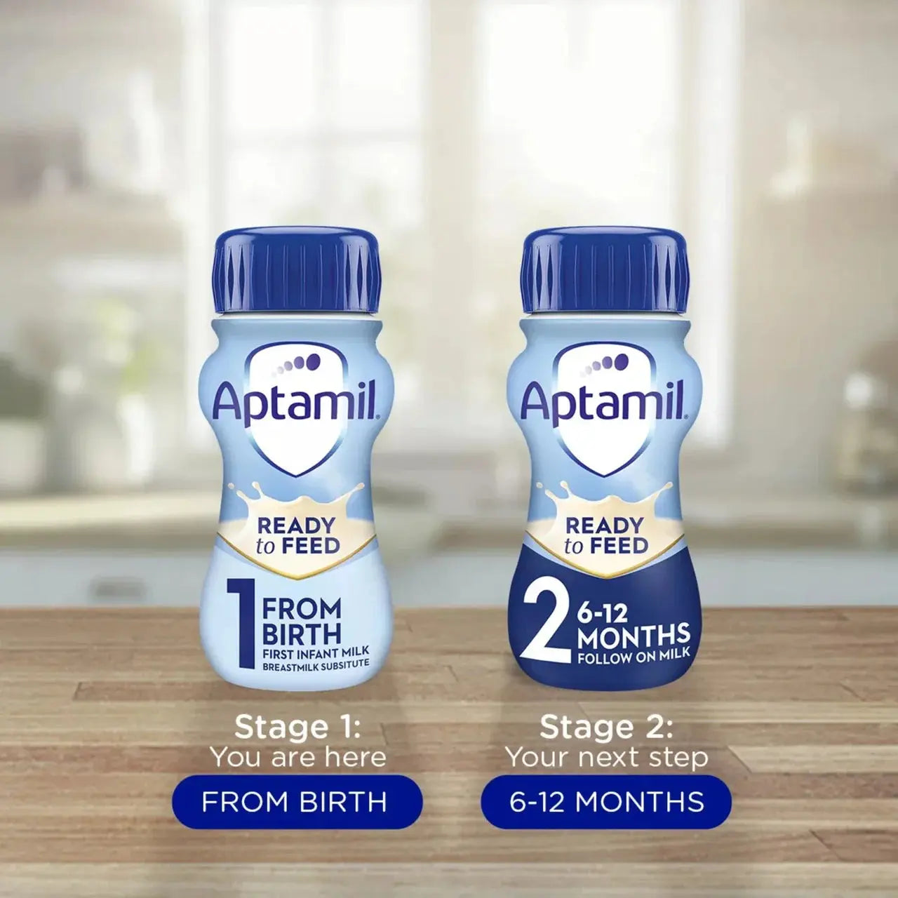 Two Aptamil Ready to Feed bottles on a wooden surface with a blurred kitchen background.
